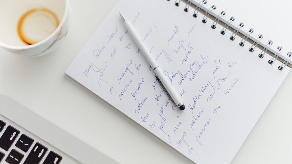 手書きのメモ帳とペン、飲みかけのコーヒー、ノートパソコンの手元。AIの下書きを自分の言葉に直す作業のイメージ