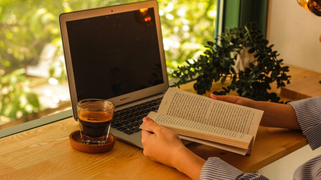 ノートパソコンの横で本を読む手元。作業をツールに任せて自分の時間を使うイメージ