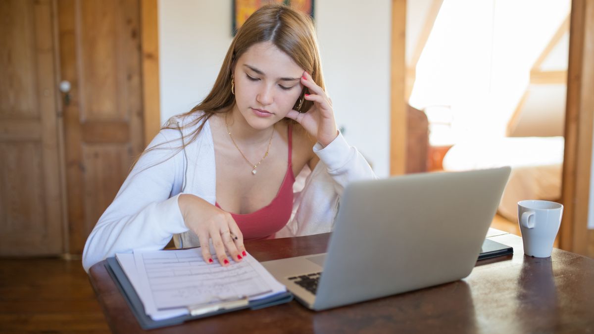 自宅の机でノートパソコンを前に、書類を指でたどりながら内容を確認している女性