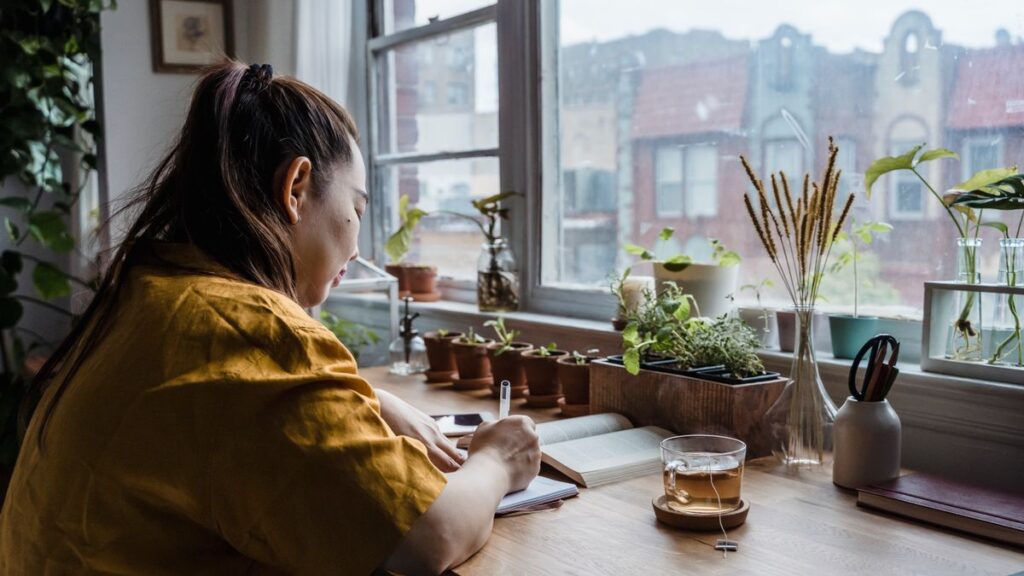 窓辺の机で観葉植物とお茶に囲まれ、ノートに書き出しをしている女性