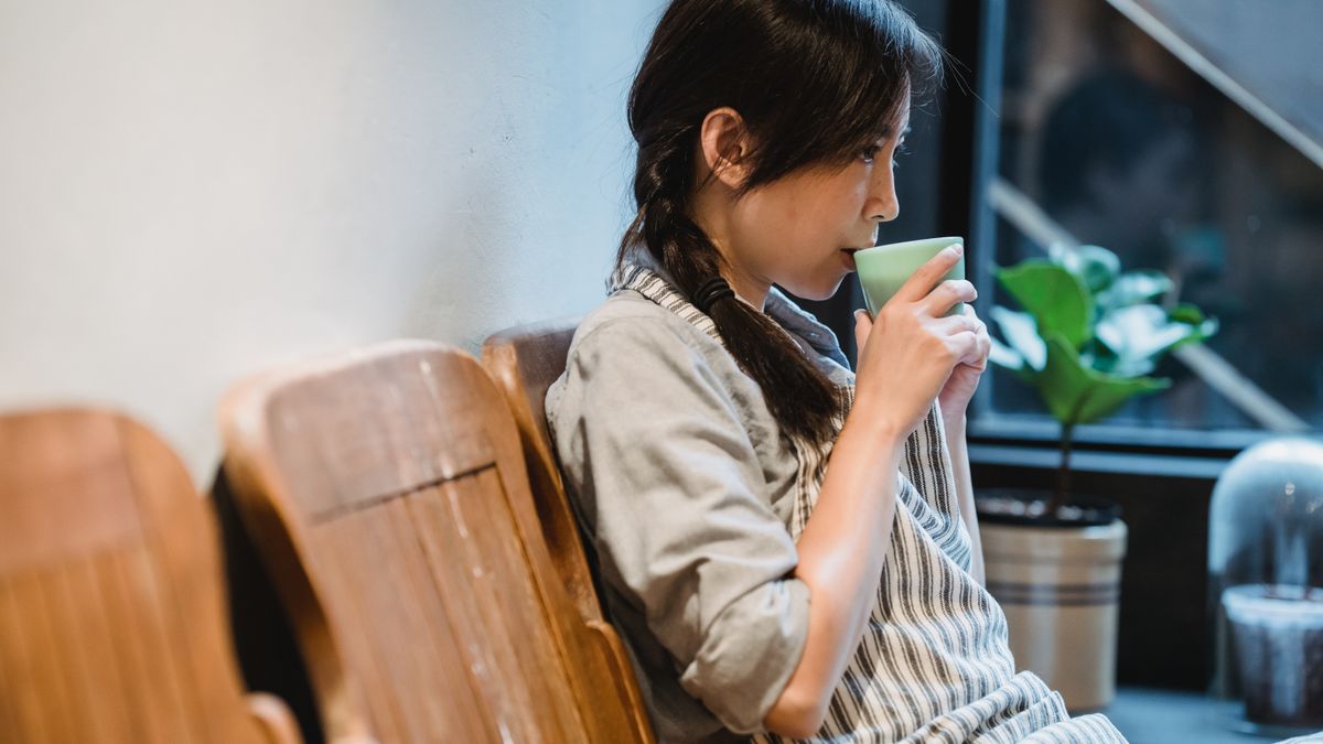窓ぎわの木の椅子に腰かけ、マグカップを両手で持って一息ついている女性