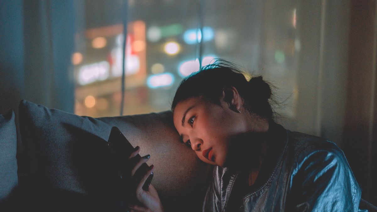 夜の部屋でクッションにもたれ、疲れた表情でスマートフォンを見ている女性