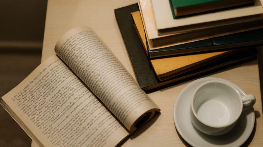 机の上に開かれた文庫本と積み重ねた本、白いコーヒーカップ。読書を習慣にするイメージ