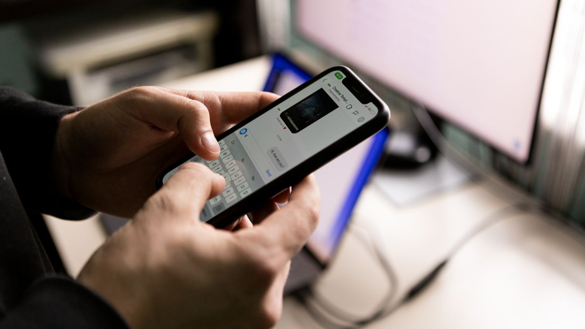 デスクのモニターの横でスマートフォンにメッセージを入力している手元